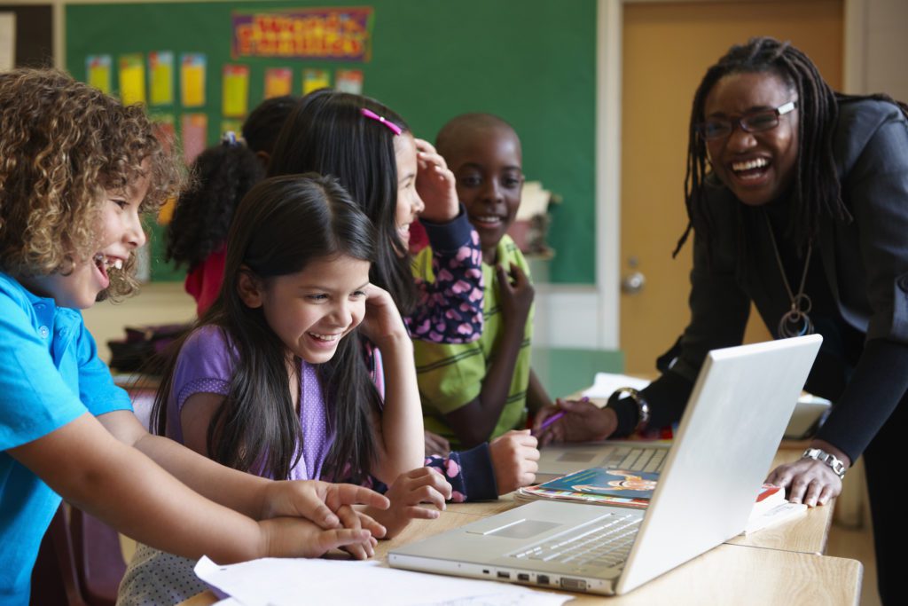 Three smiling children in front of a silver laptop and a smiling teacher in the background.